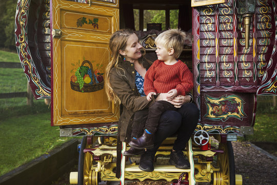 A woman and a small boy seated on the steps of a bow top caravan painted in a traditional style.