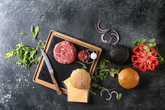 Ingredients for cooking hamburger. Meat beef burger board, cheese, ketchup sauce, tomato, black and white buns, arugula salad over dark texture background. Top view with space. Homemade fast food