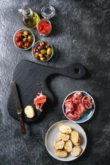 Ingredients for making tapas or bruschetta. Crusty bread, ham prosciutto, sun dried tomatoes, olive oil, olives, pepper with wooden serving board over dark texture background. Top view with space