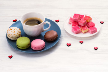 A cup of coffee with french colorful macaroons, marmalade on a white wooden table, decorated with hearts. St. Valentine's Day.