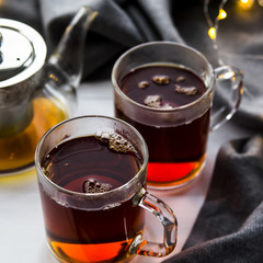 Black transparent teapot and cups with black tea on a wooden table next to a gray scarf and lights. Winter home concept