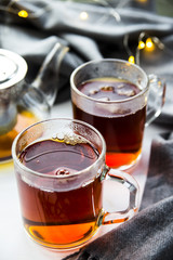 Black transparent teapot and cups with black tea on a wooden table next to a gray scarf and lights. Winter home concept