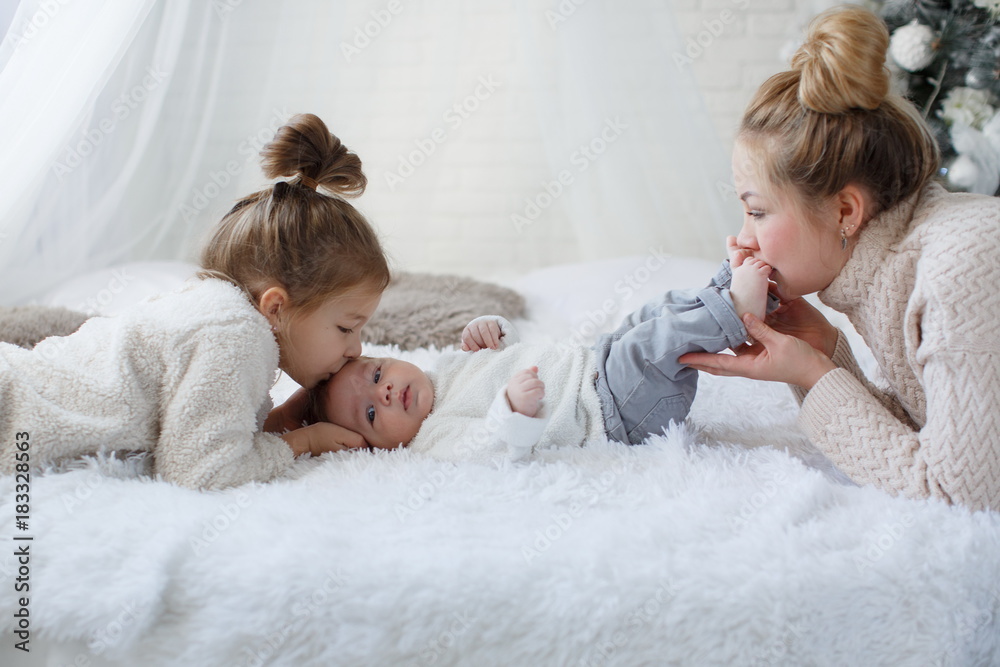 Wall mural Happy woman,cheerful mother and little children,a girl and a newborn boy sitting on a fluffy white blanket beside the Christmas festive green Christmas tree with white toys,snowflakes,glowing lights