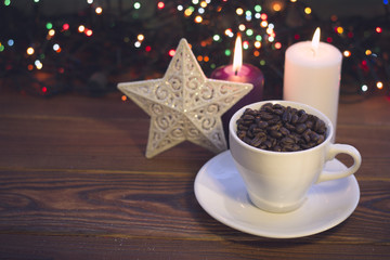 A romantic festive still life with a white cup filled with coffee beans, a white patterned toy star and purple and white burning candles on a rustic wooden table. Dark blurred bokeh background