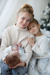 Happy woman,cheerful mother and little children,a girl and a newborn boy sitting on a fluffy white blanket beside the Christmas festive green Christmas tree with white toys,snowflakes,glowing lights