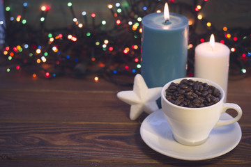 A romantic festive still life with a white cup filled with coffee beans, a white Christmas-tree toy star and blue and white burning candles on a rustic wooden table. Dark blurred bokeh background