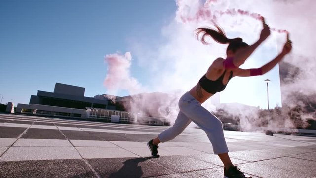 Young woman with smoke grenade jumping high performing a flashkick outdoors. Female with smoke sticks practicing tricking over urban city space.