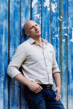 portrait of bald handsome man at natural shirt and jeans, boods. model making pose at blue wood baghround. dont look at camera. summer mood at spring time