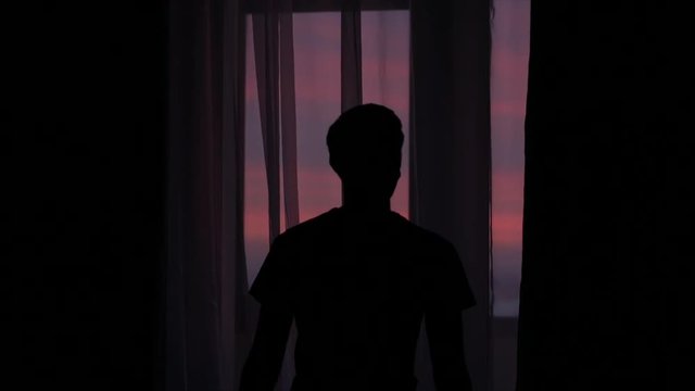 silhouette of a young man coming to a window and closing curtains against a window with a colorful sunset