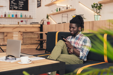 Fototapeta na wymiar cheerful african american man using smartphone and laptop in coffee shop