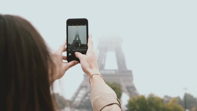 Young beautiful woman walking in Paris, France and taking photos of Eiffel tower in fog on smartphone or mobile phone.