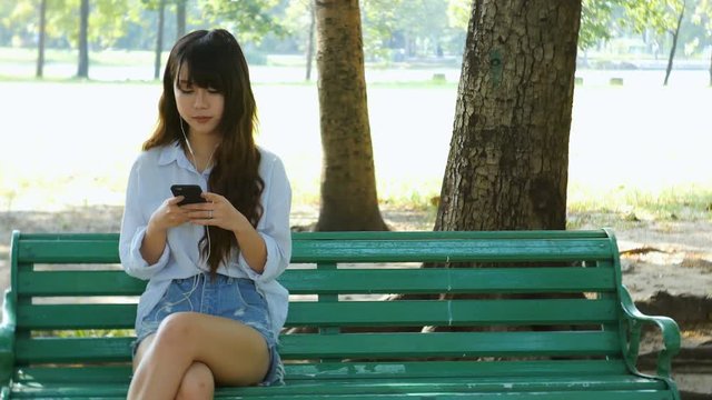Cute woman is reading pleasant text message on mobile phone while sitting in the park in warm spring day,gorgeous female is listening to music in headphones and searching information on cell telephone