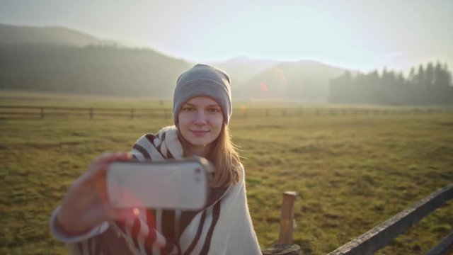 Young Woman is Taking Selfie on Front Camera of Smart Phone Outdoors with Beautiful Landscape Background at Sunset