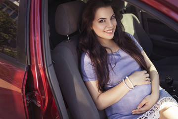 Obraz na płótnie Canvas Beautiful brunette caucasian pregnant woman on summer walk outdoors, holding her belly in a soft, sensual, maternity way. In casual jeans dress and bride hairstyle sitting in a red car. Copy space