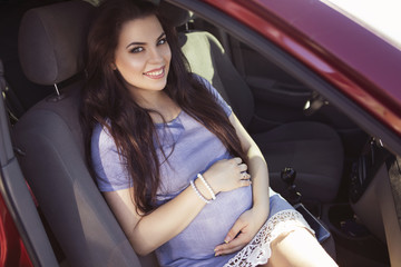 Fototapeta na wymiar Beautiful brunette caucasian pregnant woman on summer walk outdoors, holding her belly in a soft, sensual, maternity way. In casual jeans dress and bride hairstyle sitting in a red car. Copy space