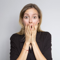 Beautiful brunette caucasian woman in dark black blouse with bright emotions standing on a neutral grey background. Her hands make a gesture. She shocked, sared