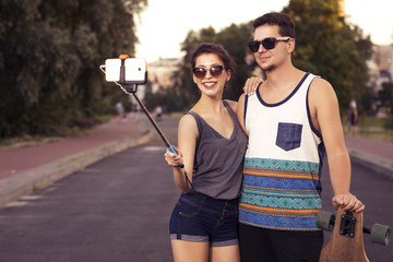 Young beautiful brunette caucasian sexy adult woman and men couple in love on a date outdoors in city, having fun skateboarding (on longboards). Making selfie on their phone. Summer weather