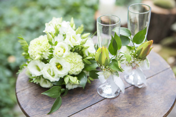 Bouquet of eustoma roses and glassess decorated with flowers lay on wooden table on a wedding ceremony. Soft day light, outdoor in garden