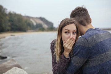 Happy thoughtful couple sitting on a rock beach near sea hugging each other in cold foggy cloudy autumn weather. Copy space