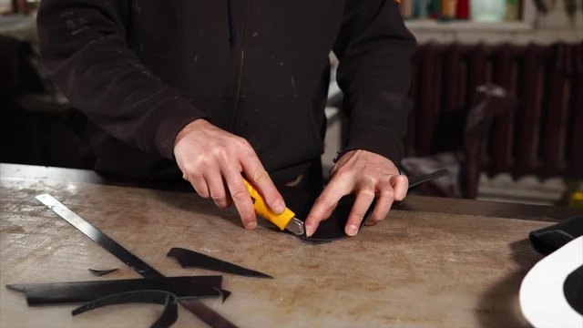 Craftsman cuts extra pieces of leather to make necessary shape. He is working with a knife on the table in a workshop.