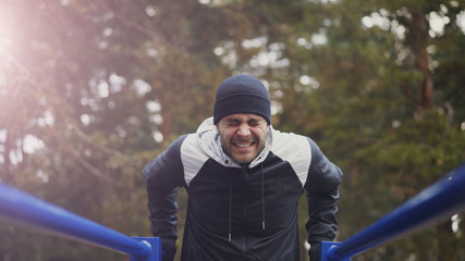 Young athlete man doing push-ups exercise on bars in winter park outdoors