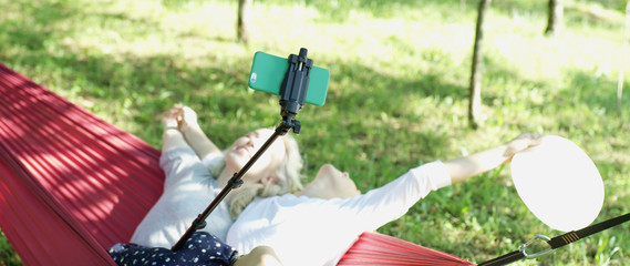 Young friends in sweaters making selfie photo with smart phone sitting together on the hammock in the garden