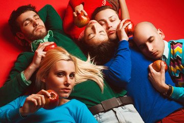 Picnic concept. Portrait of four stylish close friends hugging, having rest, holding red apples and lying on red background. Guys having fun. Hipster style. Studio shot