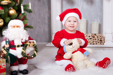 Christmas or New year celebration. Little girl in red dress and santa hat with bear toy sitting on the floor near the Christmas tree with xmas gifts. A fireplace with christmas stocking on background