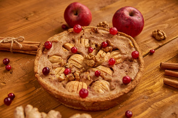 Tasty apple pie with fresh red cranberries and walnuts decorated with two apples, ginger and sticks of cinnamon. Just backed apple pie ready to eat on wooden brown background. Side view