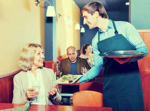 Positive waiter bringing order to smiling mature female