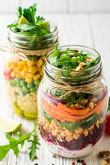 Healthy homemade salads with chickpeas, bulgur and vegetables in mason jars on white wooden background.