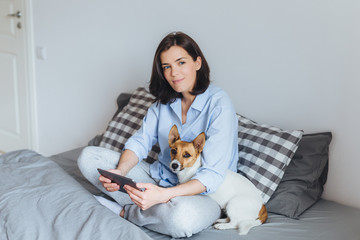 Dreamy brunette female model in pyjamas sits crossed legs on comfortable bed in bedroom, holds...