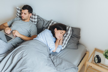 Pretty female sleeps in bed, sees pleasant dreams, while her husband works on laptop computer, prepares report, tries not to interrupt his wife sleep. Family couple in bedroom. Rest concept.