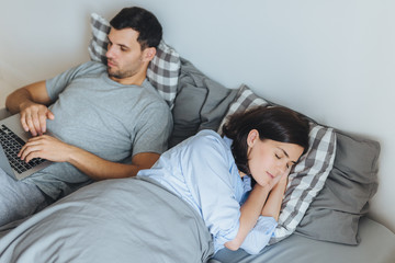 Busy male lies in bed, works on laptop computer, has serious expression while his wife has healthy sleep, being together in bedroom. Two people in bed. People, family, home concept
