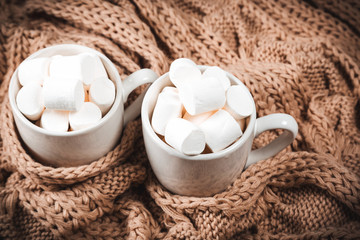 Fototapeta na wymiar Cocoa or coffee with marshmallows in a white cup with a brown knitted winter scarf. Beautiful christmas background