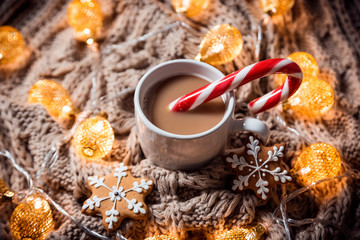 Christmas background coffee in a white cup and gingerbread snowflakes with candy on a brown knitted winter scarf and a glowing golden garland. Beautiful home Holiday drink