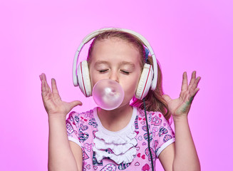 little girl with headphones blowing up a bubble of gum on a pink background
