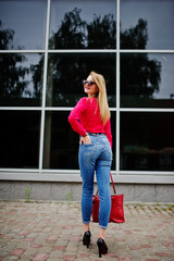 Portrait of a fabulous young woman in red blouse and jeans posing with her handbag and sunglasses outside the shopping mall on glass background.