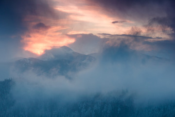 Landscape of dramatic sunset in the winter mountain. Wooded hills covered with snow, fog rising from valleys, colorful cloudy sky - this is impressive picture.
