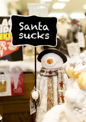 Ornamental snowman wearing a scarf, coat and a top hat holding up a Santa Sucks sign in a Christmas shop