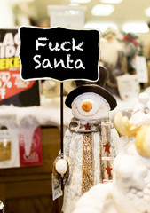 Ornamental snowman wearing a scarf, coat and a top hat holding up a fuck Santa sign in a Christmas shop