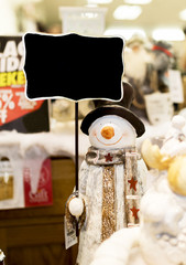 Ornamental snowman wearing a scarf, coat and a top hat holding up a blank sign in a Christmas shop