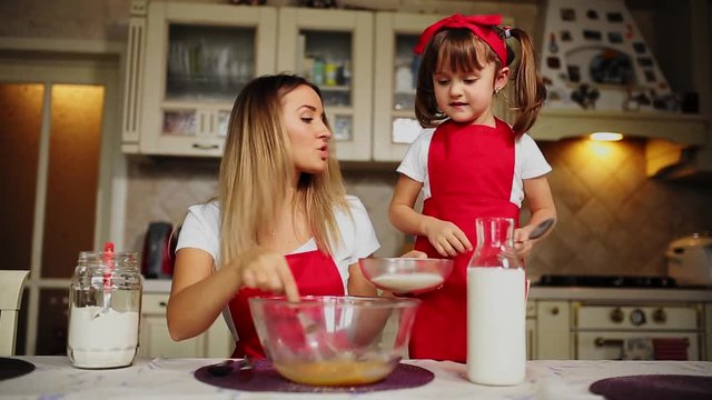 Beautiful young mother helping her little daughter along to cook cake in red aprons. Pour the flour into a bowl and beat the batter to make a cake in the kitchen.