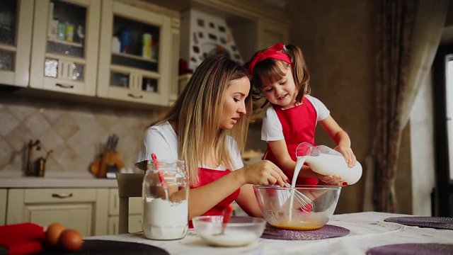 Beautiful young mother helping her little daughter along to cook cake in red aprons. Pour the milk and stir the batter together in the kitchen.