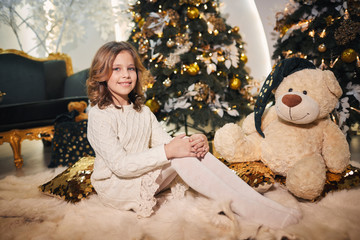 beautiful baby in a beautiful warm white sweater holds newlyweds gifts near a tree with toys, garlands and lights
