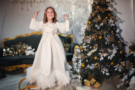 little girl who dreams of being a princess standing next to a Christmas tree dressed in a white luxurious dress and a silver crown