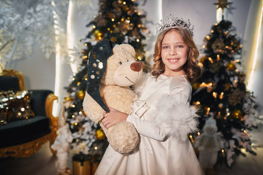 a little child dressed in a costume of a snow queen posing near a Christmas tree with toys, garlands and flashlights