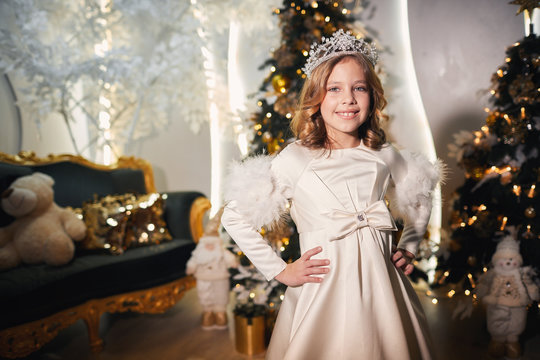 a small queen in a long and lush white dress with a silver crown on her head sitting on a magnificent green sofa of a white Christmas tree and smiles