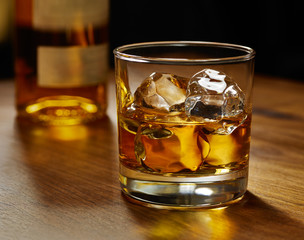 Glass of whiskey on ice with bottle on wood bar
