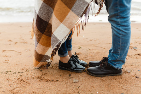 View Of Feet Of Couple In Love Standing On The Shore Of A Natural Beach Wrapped Up Sharing A Blanket On A Cold Winter Rest. A Guy And A Girl Are Traveling Lifestyle, Tranquility And Contemplation.